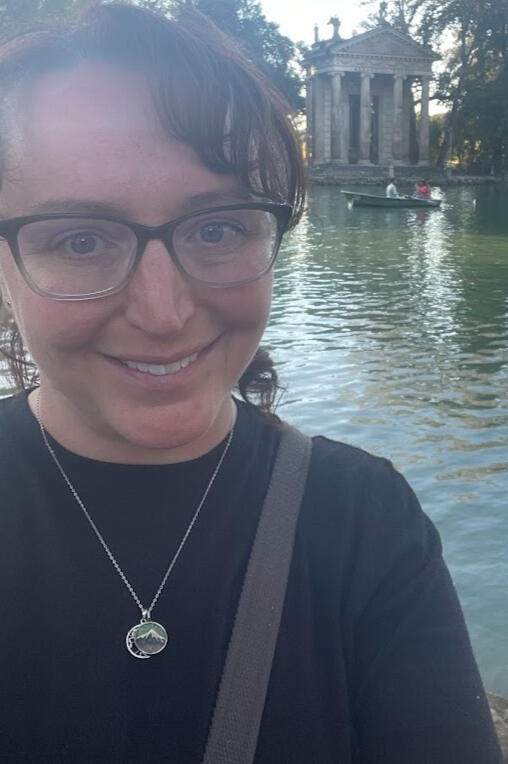 Woman smiling at camera in a black shirt with a pond and column building behind her, she is on the left side of the image